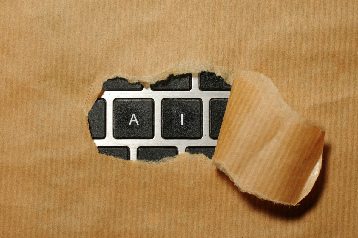 The image is a creative and surreal visual metaphor. It depicts a laptop keyboard partially obscured by a piece of paper that has an opening in the middle revealing a white computer keyboard with black keys, which is the focal point of the photo. There are two revealed keys, 'A' and 'I' The background appears to be a cardboard surface with a hole cut out, allowing the viewer to see through it and focus on the keyboard, creating a peek-a-boo effect. This artistic composition suggests themes of secrecy, information technology, or perhaps even the idea of 'keying in' or accessing something hidden beneath the surface. The use of everyday objects like a laptop and cardboard in this way adds an element of whimsy and intrigue to the image.
