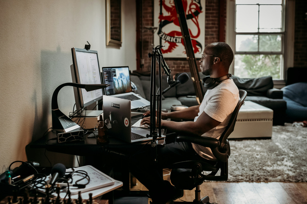 In the center of the room, a man is seated at a desk, engrossed in his work on a computer. He's dressed casually in a white t-shirt and blue jeans, suggesting a relaxed atmosphere. The computer equipment, including a monitor, keyboard, and mouse, suggests he might be involved in content creation or broadcasting, as indicated by the microphone in front of him. The room itself is spacious, with high ceilings that house exposed ductwork, adding an industrial touch to the space. The walls are adorned with artwork, including a dragon-themed piece on the left and a red dragon on the right, adding color to the otherwise neutral tones of the room. A large window in the background allows natural light to flood the room, creating a warm and inviting ambiance. On either side of the desk are two comfortable couches, one on each end of the room, offering alternative seating options. The floor is covered with tiles that reflect some of the room's features, enhancing the sense of spaciousness. Despite the numerous elements in the room, everything seems to be arranged harmoniously, creating a functional and stylish workspace.