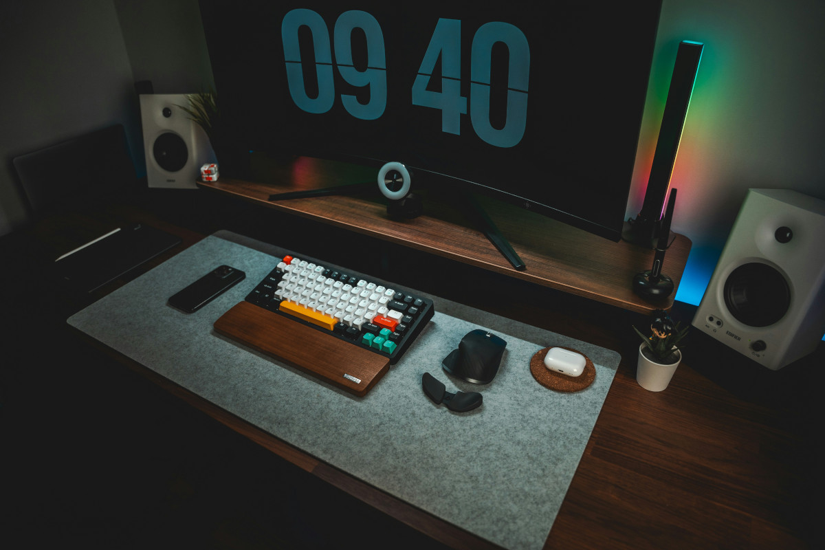 A well-organized computer desk with a monitor displaying the time '09:40'. The desk has a keyboard, mouse, and various  other electronic devices. The foreground features a keyboard and mouse, the keyboard has a colourful array of keys.