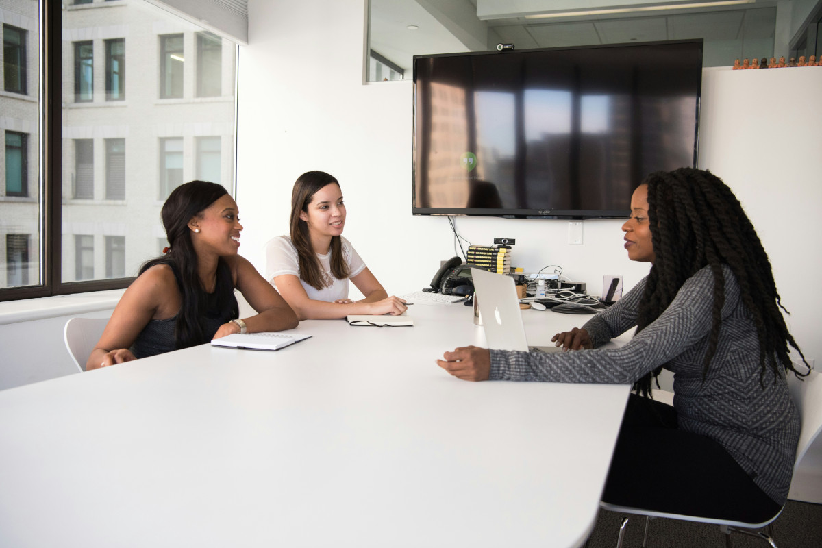 This is a photograph showing three individuals in an indoor office setting. All three individuals are seated, two on the left and one on the right. Those on the left are looking at the person on the right, whilst the person on the right is looking at one of the people on the left. All are engaged in conversation. The person on the right has a laptop open in front of them. The room has a modern design with a large window that provides natural light, contributing to a bright atmosphere. There's a TV mounted on the wall in the background. The people are dressed professionally, suggesting they are in a formal work environment.