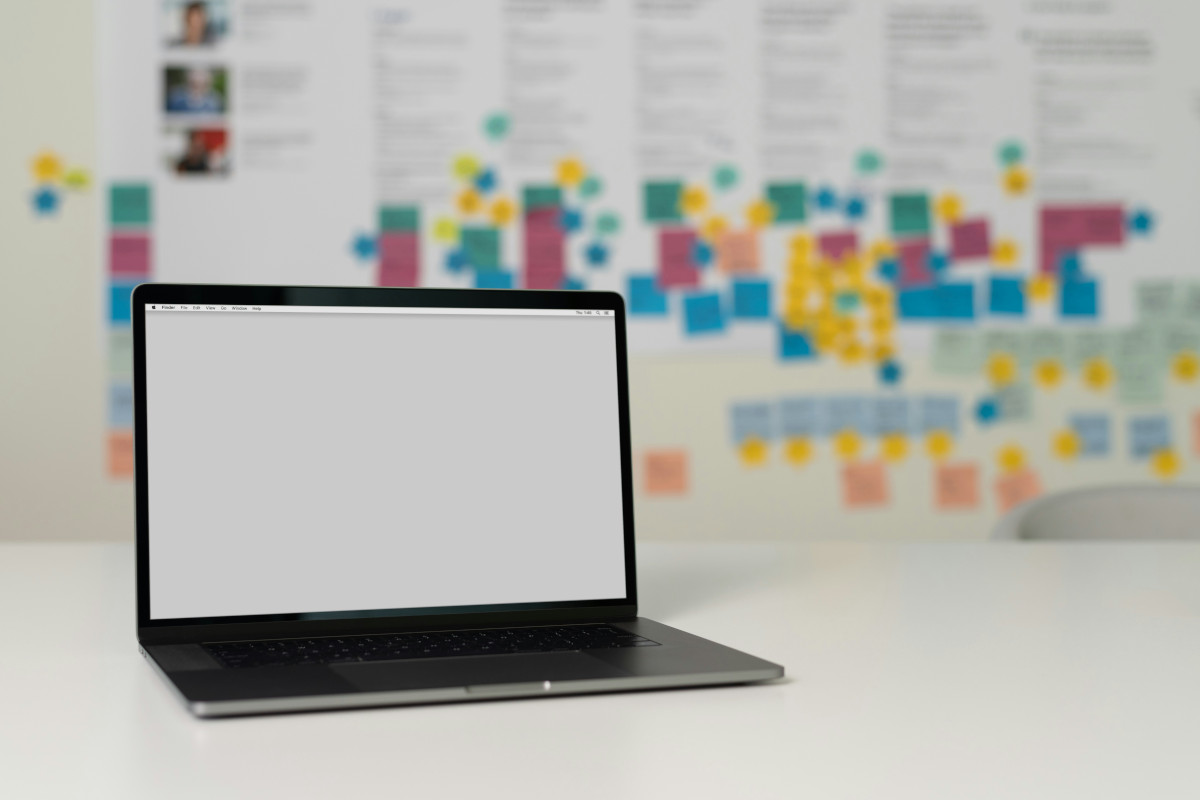 The image shows a laptop computer sitting on a table with a white surface. The laptop is open and displays a blank screen. Behind the laptop, there's a wall with various post-it notes and diagrams attached to it, suggesting an organized workspace or brainstorming area. These notes are clustered and arranged in no particular order. The room has a modern, professional ambiance. There are no visible people or distinctive features that allow for specific identification of the location or event. The focus of the image is on the laptop, which appears to be the main subject.