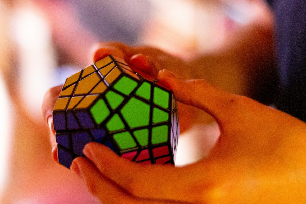 In the image, a person is holding up a Rubik's Cube in their hands. The cube has multicolored pieces, with each side featuring different colors. The person appears to be indoors with warm lighting and there are out-of-focus objects in the background that suggest a casual, home environment. 