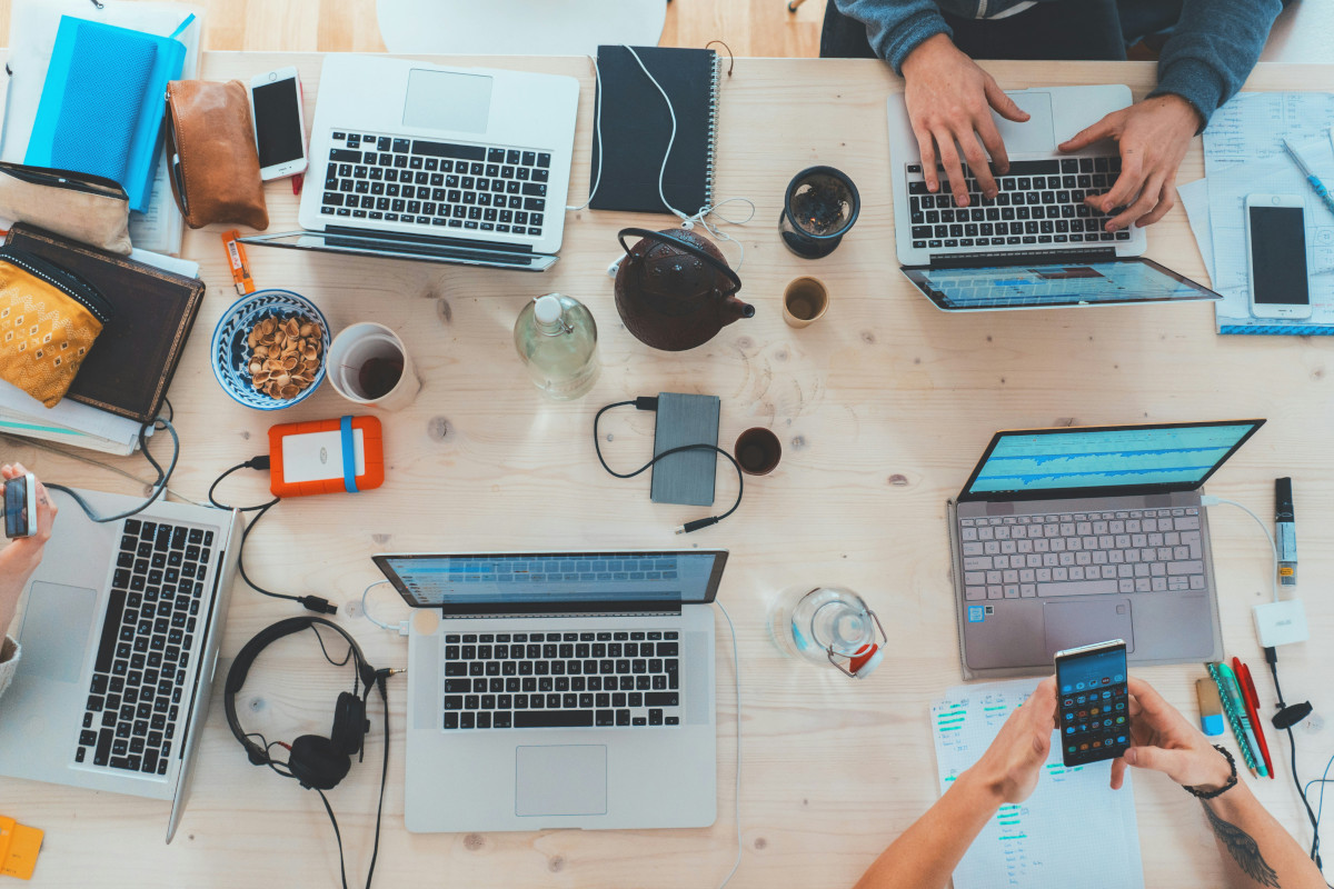 The image depicts a collaborative work environment, with several people engaged in activities around laptops and various other office equipment. There are four individuals present in the scene; one is working on a laptop, while others are observing or interacting with their devices. On the table, there are multiple cups of coffee, suggesting that they have been working for some time. The table itself has several personal items like bottles, a book, and a notebook on it. The laptops are open, indicating active use, and some peripherals like keyboards are visible near them. There is a chair in the scene, likely used by one of the individuals. The background shows a cozy and informal setting with a wooden table and a wall adorned with what appears to be artwork or decorations. The overall atmosphere is casual yet focused, indicative of a modern, flexible workspace designed for creative professionals or teams. 