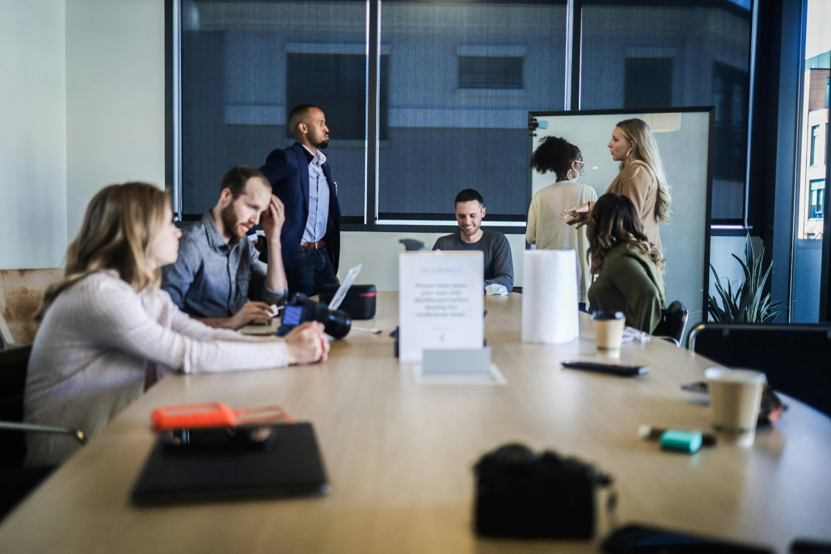The image is a photograph that shows an indoor setting where a group of people are sitting around a large table. They appear to be engaged in a meeting or discussion, as suggested by their focused expressions and the presence of what looks like a presentation or notes on the table. There are multiple laptops open in front of some individuals, indicating a working environment. The background is blurred, but it seems to be an office space with modern decor, including plants, which contribute to a casual yet professional atmosphere. There's no text visible in the image that provides context or information about the people or event taking place.
