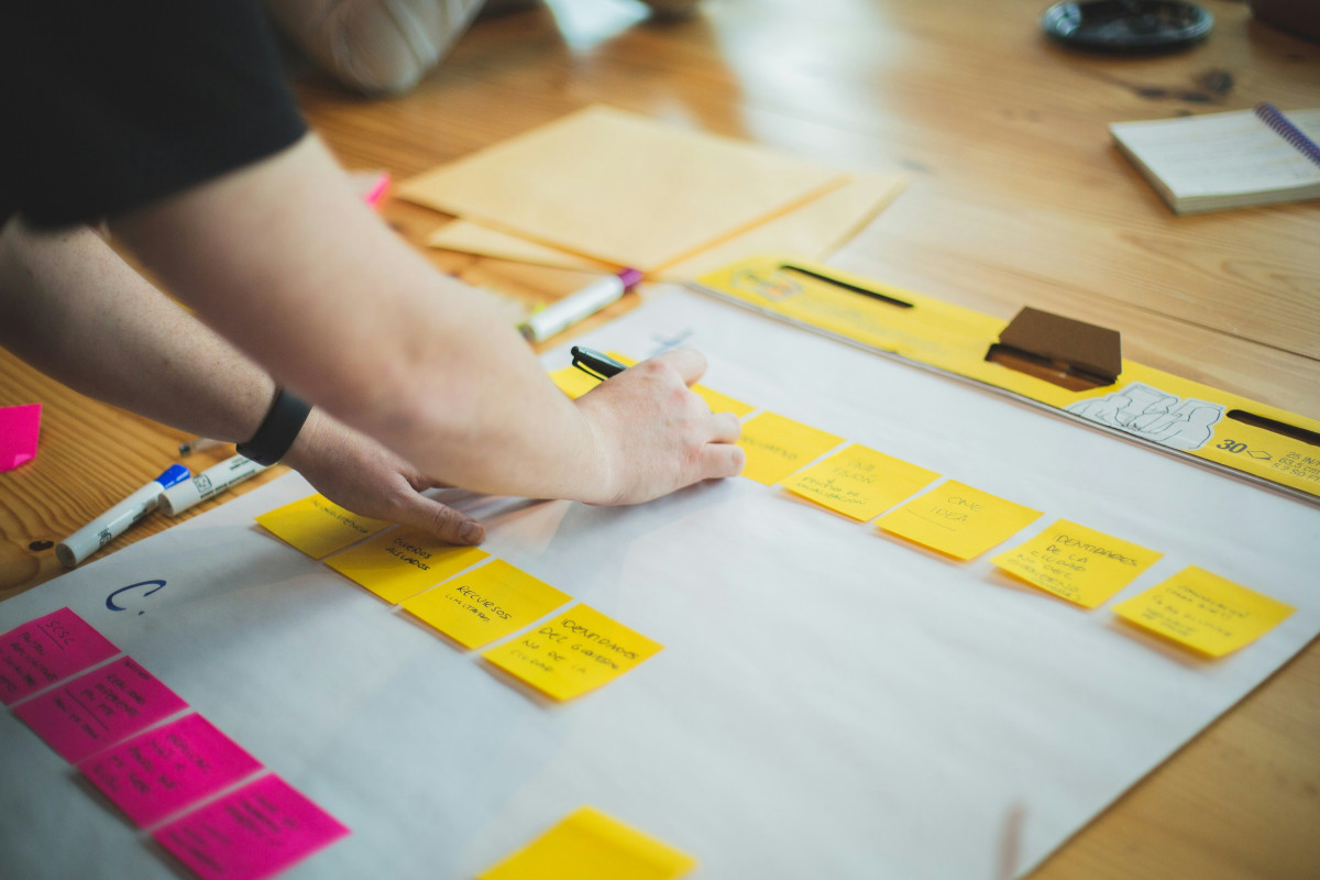 A person is working on a project at a wooden table. The individual is focused on an array of sticky notes attached to a chart, which are arranged in a horizontal line. The table also holds some pens and notepads. The environment suggests a planning or brainstorming session, possibly related to a work or creative project.