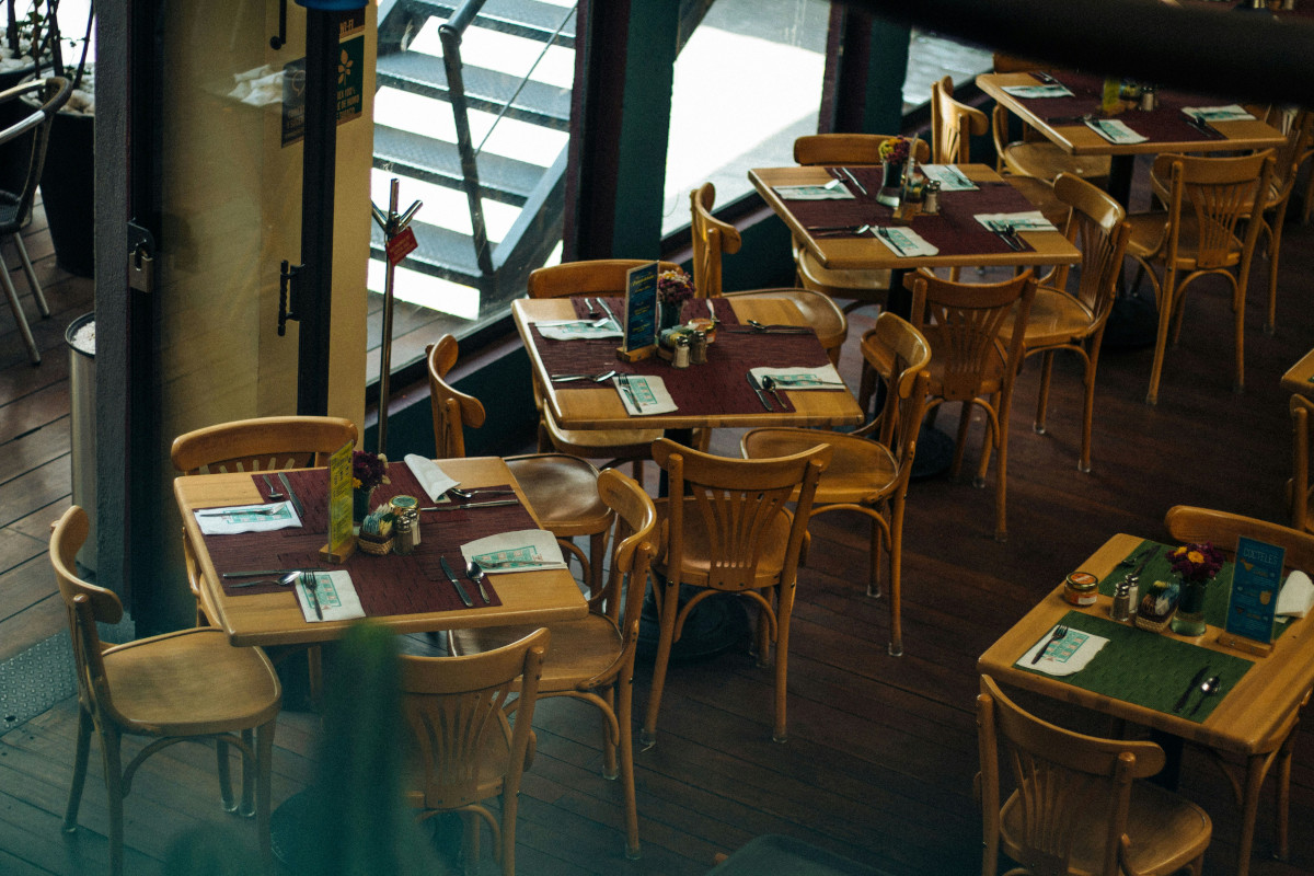 This is an image of a well-arranged dining area inside a restaurant. There are several wooden tables with chairs, set up neatly and covered with place settings including white plates and napkins, indicating that the establishment is prepared for service. The floor is made of wood planks, adding to the warm ambiance of the room. On the right side of the photo, there's a counter with a glass top, possibly serving as a bar or additional dining area. The ceiling has an exposed structure with beams and ductwork, contributing to the industrial chic aesthetic. The overall setting suggests a casual yet stylish dining environment.