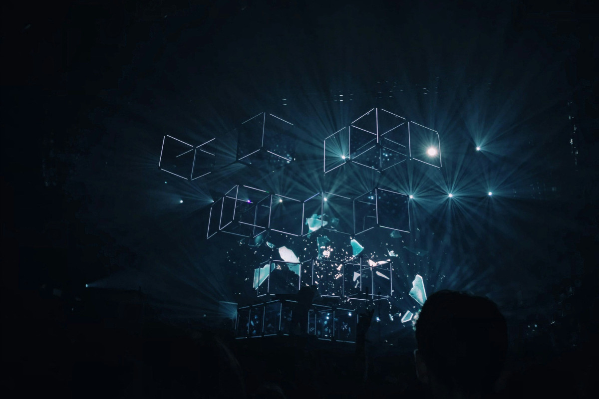 The image is a photograph taken at an indoor venue, likely a concert or live performance. The stage features a colourful array of lights and geometric shapes creating a  digital, glowing effect that resembles futuristic technology or cubes. The light colors used in the design are blue, green, and white. There are several beams of light  emanating from these shapes, contributing to a dynamic and vibrant atmosphere. In the background, there is a crowd of people who appear to be spectators. They are partially visible with their backs towards the camera, suggesting they are facing the  stage. The lighting in the venue casts shadows on the walls and ceiling, indicating the presence of spotlights or directional lights focused on the performance area. There are no texts visible within the image. The style of the photograph captures the energy of a live music event and conveys a sense of movement and light through  colourful illumination and architectural elements.