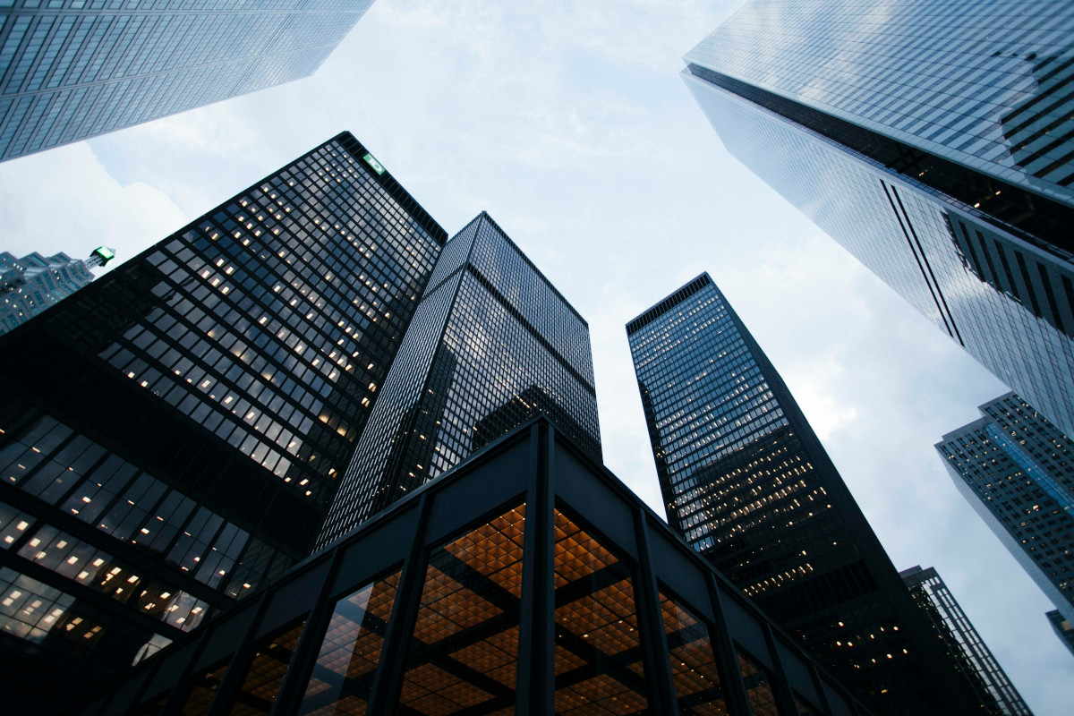 The image depicts a close-up view of the facade of a tall, modern skyscraper during dusk or early morning. The building is architecturally designed with a reflective glass exterior that suggests contemporary style. There are lights illuminating the facade, creating a warm glow against the cooler tones in the background. The sky is overcast with gray clouds, hinting at an urban environment. In the foreground, there's a clear reflection of the building on what appears to be a glass surface, possibly another high-rise building nearby. At the bottom of the image, you can see two rows of illuminated windows that span across the height of the building. These windows are neatly spaced and evenly lit, indicating that they could serve as offices or meeting rooms within the building. The photo is taken from a low angle, looking up towards the skyscraper. This perspective emphasizes its height and grandeur, while also capturing elements of street level, such as the reflection on the glass surface, which suggests another building is in front of it. The style of the image is realistic with a slight focus blur, which brings attention to the central object—the skyscraper.