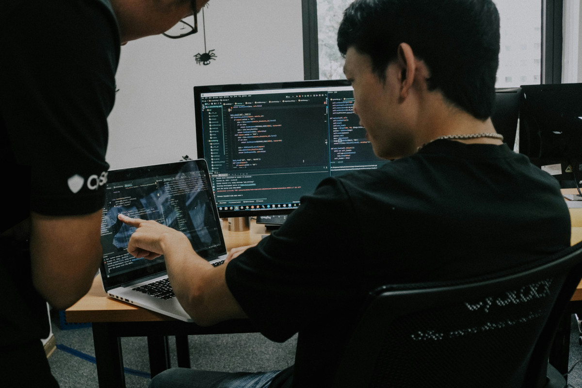 The image depicts a man sitting at a desk with his attention focused on the screen of a laptop computer. He appears to be engaged in some work or activity, possibly related to software development or coding given the open source code editor displayed on the laptop screen. Behind him, another person is visible from the waist up; they are standing and facing away from the camera, suggesting an interaction between the two individuals, potentially discussing a project or sharing information. The setting seems to be an office or workspace with a professional ambiance, indicated by the tidy desk and the presence of technical equipment.