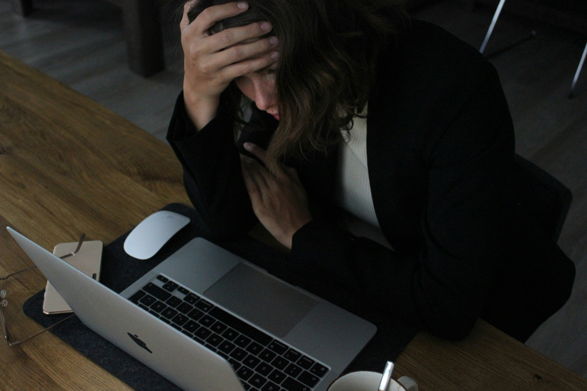 This is a photograph of a person sitting at a desk, with their head resting on one hand. The individual appears to be in a state of frustration or exhaustion, as suggested by their facial expression and posture. In front of the person, there is an open laptop, we cannot see what it says. They have discarded their glasses on top of an overturned iPhone, indicating a stressful situation. The overall mood of the image conveys stress or overwhelm, commonly associated with high-pressure jobs, especially in technology fields where multitasking and information overload are prevalent. The focus of the photograph is on the person's head and upper body, making it a close-up shot capturing an intense moment.