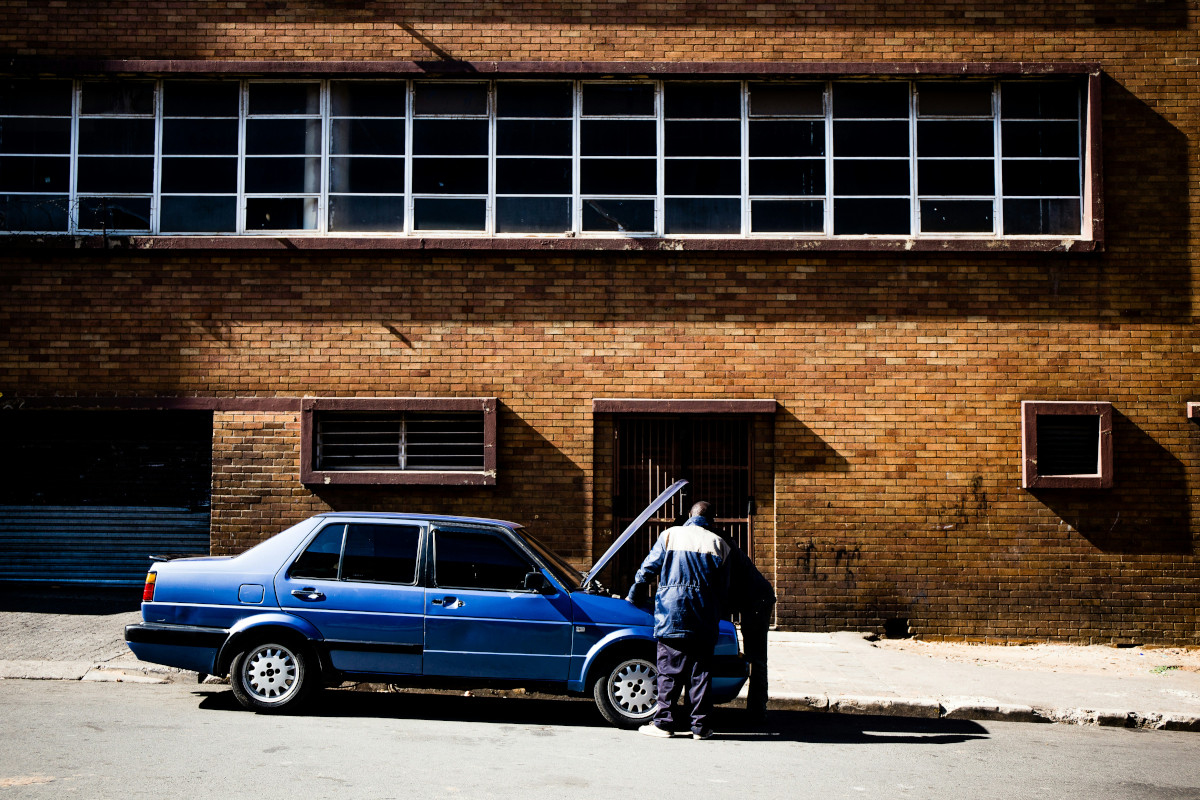 The image shows a scene featuring a person standing next to an old blue car parked on the side of the street. The car is facing away from us, with the driver's side visible. Behind the car and the person stands an imposing brick building with large windows on both sides, giving it a somewhat dilapidated appearance. There are signs of vandalism or urban decay, such as graffiti or marks on the walls. The setting suggests an urban environment during the daytime, given the shadows visible on the ground.