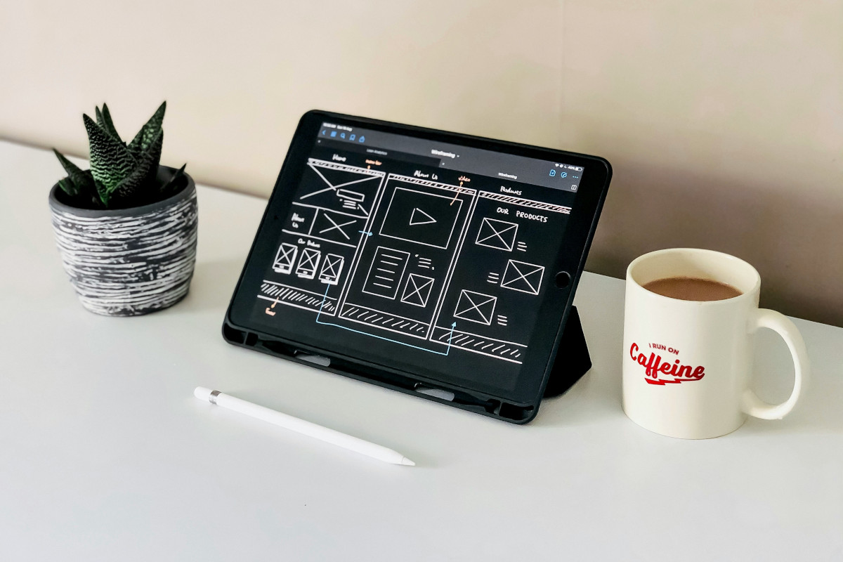 The image is a photograph of a tabletop with various items on it. There's an iPad open to what appears to be a drawing application, showing a digital sketch or design with several layers and tools visible on the screen. In front of the tablet, there are two white mugs that say 'Caffeine,' which suggests they might be for coffee. To the right of the table, partially out of frame, there is a black plant with spiky leaves. The background is neutral, focusing attention on the items in the foreground.