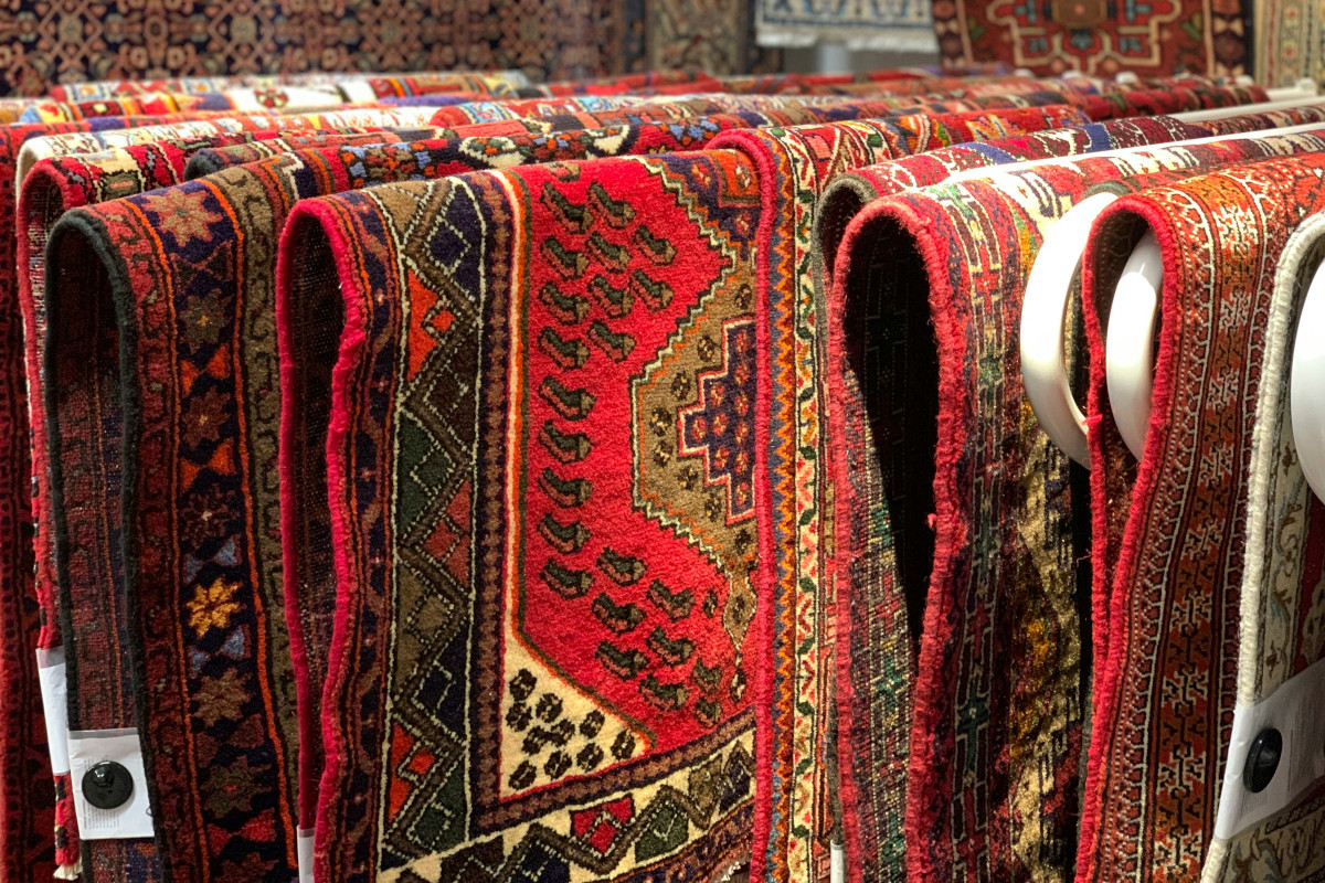 A number of multi-coloured carpets are arranged on a rack. They are predominantly red with yellow, bronze, and gold motifs.