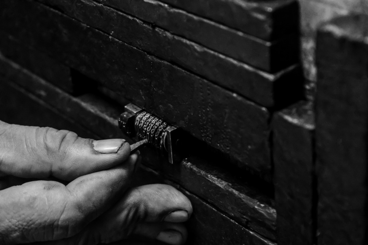 A monochrome close up image of a person picking lock. the lock is in the centre of the image, and the person's left hand can be seen reaching in from the left-hand side of the frame.