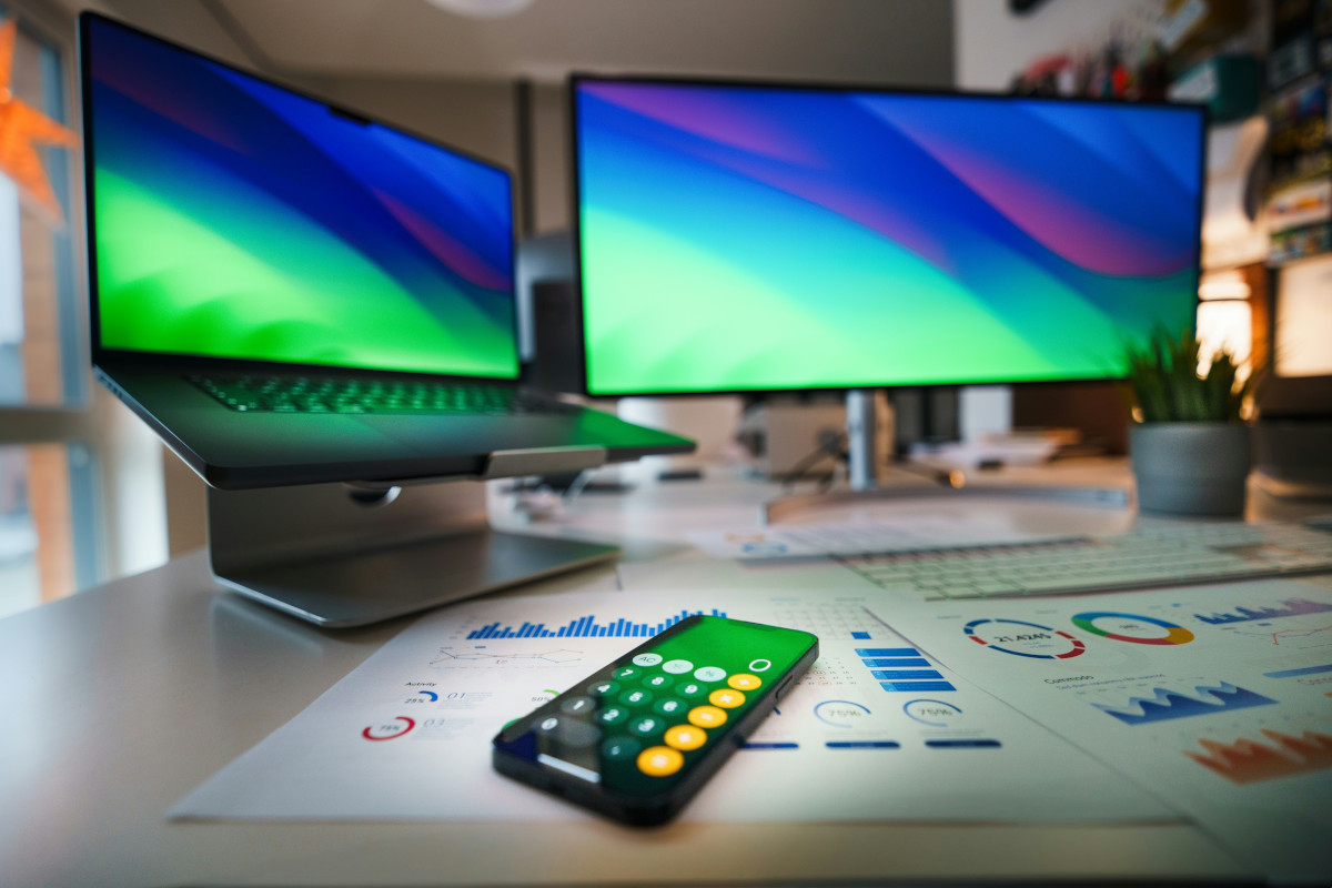 This is a photograph showcasing a modern office setup. There are two computer monitors on adjustable stands, displaying colourful wallpapers with abstract designs in hues of green, blue, and purple. The screen on the left-hand side of the image is actually a smaller laptop screen.
Below the monitors, there's a sleek smartphone with the calculator app open. In front of the monitors on a desk is a potted plant, and some documents showing various charts and graphs indicating this is the desk of someone who works in finance. The documents are out of focus, and we cannot see any fine detail on them.