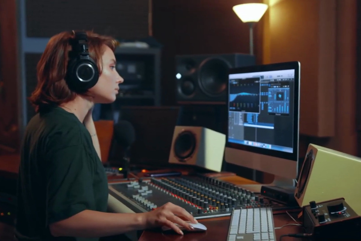 The image shows a person sitting at a computer workstation. They are wearing headphones and appear to be engaged in some audio or music production tasks, as suggested by the presence of audio equipment and a software interface on the screen. There is a monitor displaying what looks like a digital audio workstation (DAW) with various panels and waveforms visible. The person is dressed casually, suggesting a relaxed or informal setting, possibly a home studio or an office within the creative industry. The background is blurred but includes elements that suggest a professional workspace, such as additional audio equipment and possibly soundproofing materials. The overall scene conveys a sense of creativity and focus on audio production.