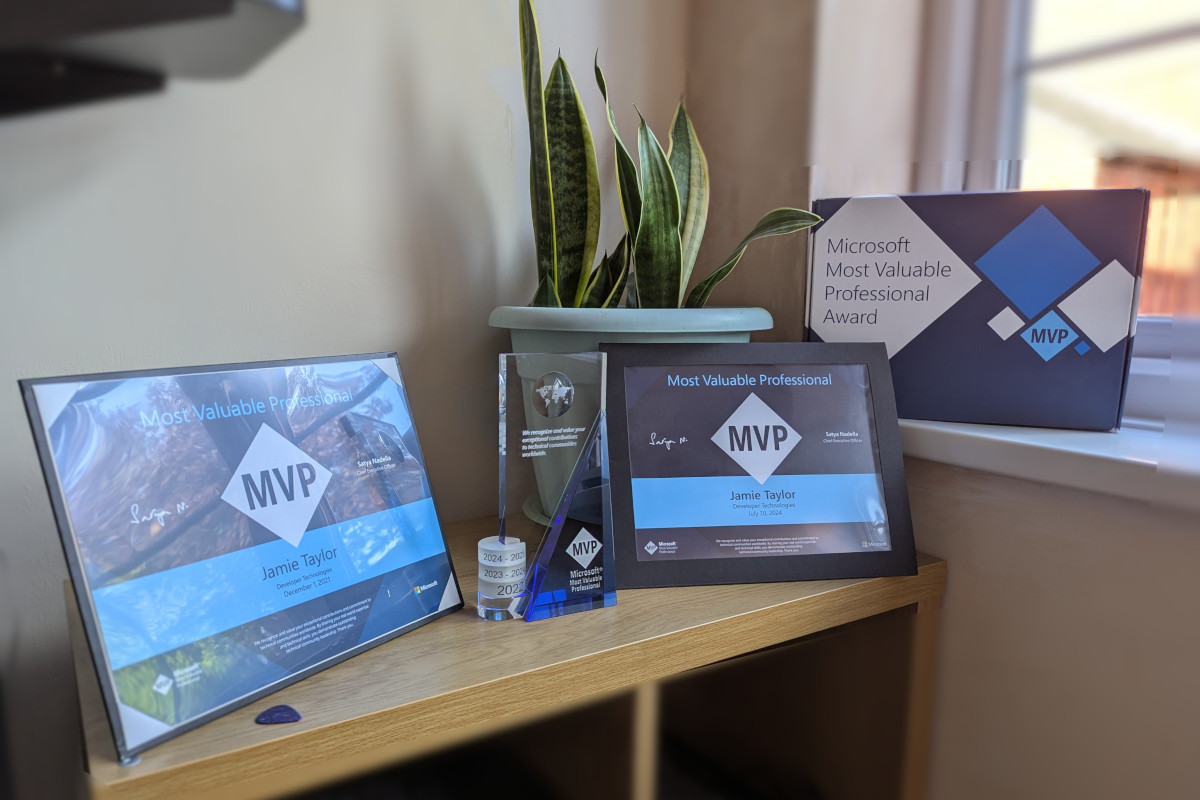 The image shows a collection of items on a surface. At the center, there's a framed certificate with visible text that reads 'MVP' in blue, suggesting it may be an award or recognition. To its left, there's a small, potted plant. On the right side of the certificate, there is a glass trophy with a blue ribbon and a gold medal base, also signifying an award or achievement. In front of these items, there are two brochures or flyers leaning against each other. The brochure on the left appears to have text, but it's not clear enough to read accurately. To the right, there is another trophy with a similar blue ribbon, placed slightly behind the first one. Behind these items, a window can be seen allowing natural light into the room. The overall setting seems to be an office or personal workspace. The style of the image is a straightforward photograph, likely taken to document the awards and their presentation.
