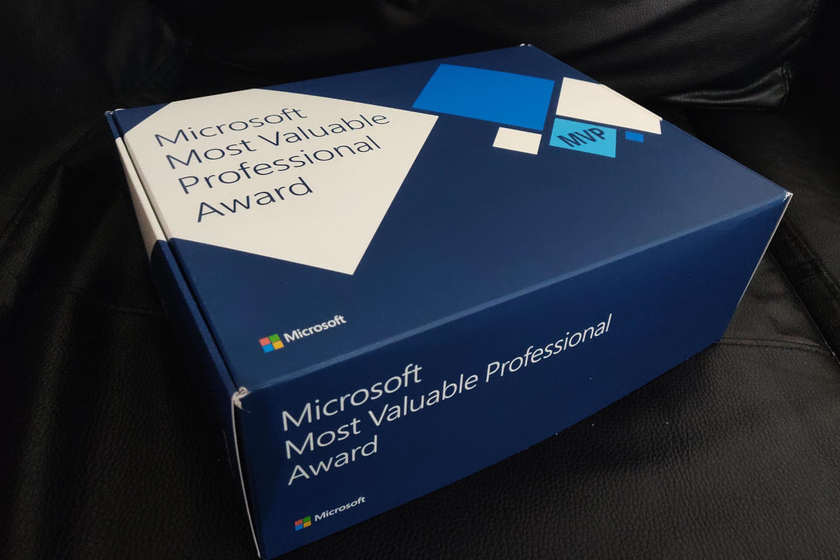 The image shows a box that is labeled 'Microsoft Most Valuable Professional Award.' The box is placed on a surface with a dark background, and there's a piece of paper next to it. The paper has the same label as the box, indicating it might be related to the award or documentation about it. The overall setting suggests a professional or office environment.