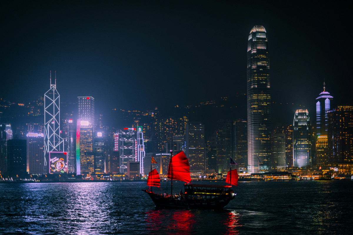 A night scene with a modern cityscape in the background. Prominent buildings and landmarks, including Hong Kong's skyline, are illuminated against the dark sky. The focus of the photo is a body of water where boats are visible, including one with red sails. In the foreground, there is a skyline silhouette against the night sky. The overall mood is peaceful and urban
