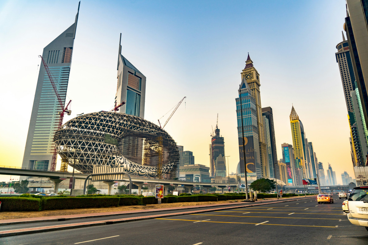 The image shows an architectural structure with modern designs and a futuristic appearance, resembling a conceptual building model. The photo captures the cityscape of a large metropolis during what appears to be sunrise or sunset, given the soft hues in the sky. The skyline includes high-rise buildings. In the foreground, there are roads with parked cars and traffic lanes, indicating the view is from an elevated vantage point or possibly through a car window due to the reflections on the glass.