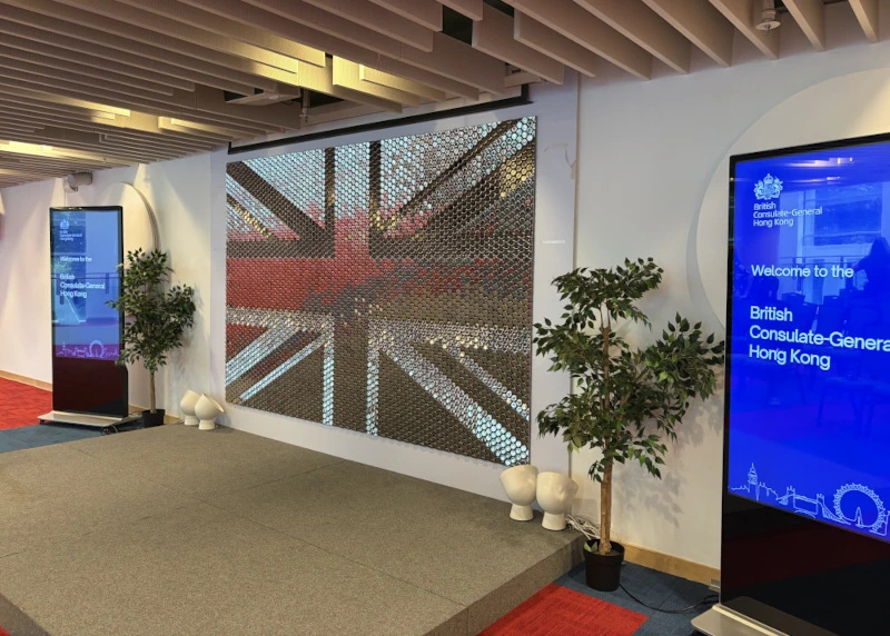 The image shows an interior view of a room, likely a lobby or exhibition space. In the foreground, there is a large projection screen displaying a reflective Union Jack flag pattern. To the right of the projection screen, there's text on a sign that reads 'Welcome to the British Consulate-General Hong kong,' with a name and title below it which are partially obscured.