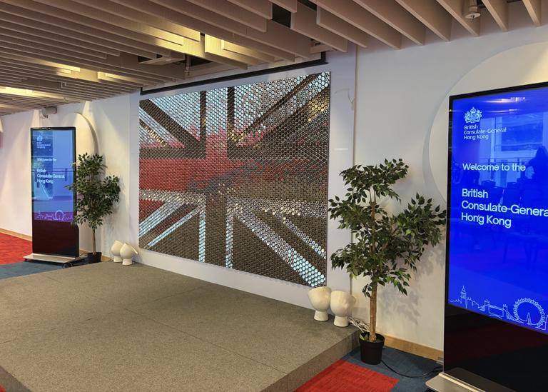 The image shows an interior view of a room, likely a lobby or exhibition space. In the foreground, there is a large projection screen displaying a reflective Union Jack flag pattern. To the right of the projection screen, there's text on a sign that reads 'Welcome to the British Consulate-General Hong kong,' with a name and title below it which are partially obscured.