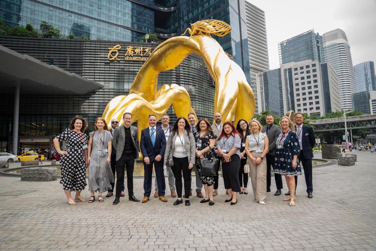 In the image, there is a group of nine individuals standing in front of a large, golden sculpture that resembles two intertwined snakes. The sculpture is prominently displayed on a pedestal and has a polished appearance. Behind the group, there is a modern building with glass walls and a signboard above it displaying Chinese characters. To the right of the group is a plaza area with a fountain, and further back, there are more buildings under a cloudy sky. The individuals in the photo appear to be dressed for a formal or professional occasion, as indicated by their attire..