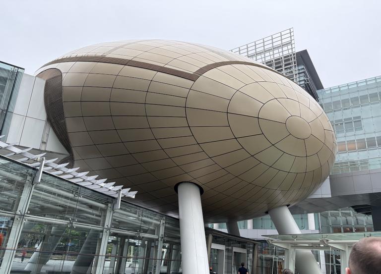 The image shows a modern architectural structure with an unusual design resembling a large, rounded, metallic object that is partially enclosed in a building. It has a reflective surface and appears to be an artwork or an installation rather than a functional part of the building. In front of this structure, there's a person taking a photo. The setting suggests it could be an outdoor public space, possibly near a train station given the architectural style and the presence of what looks like a platform with signs on it. The sky is overcast, indicating the photo was taken during daylight hours.