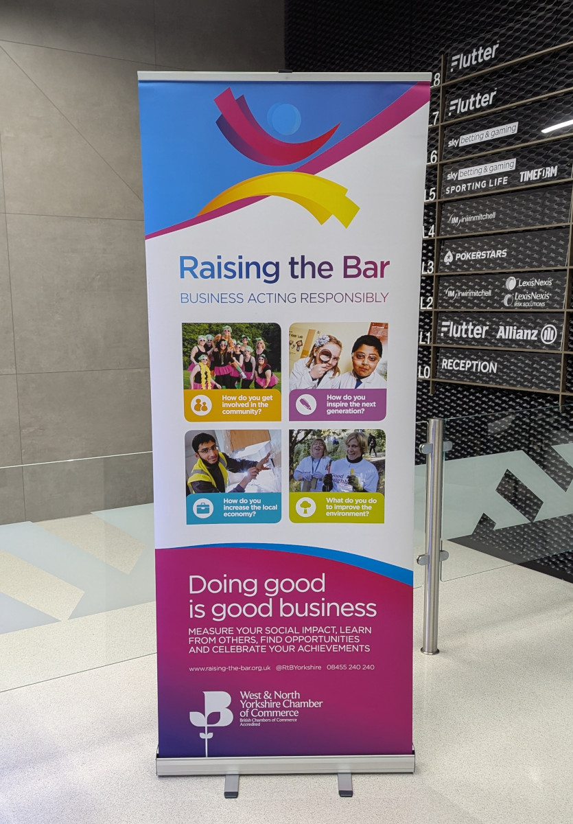 A banner stands on a tripod in front of an indoor building, advertising a charity event. The banner features various colourful graphics and text related to the event. The main message ‘RAISING THE BAR’ is prominent, along with additional information like ‘DOING GOOD IS GOOD BUSINESS.’