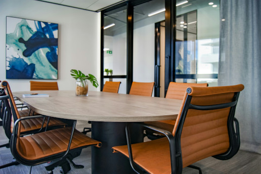 This is a photograph of an interior office space. In the foreground, there is a wooden conference table surrounded by five modern chairs with high backs and metal legs. The table appears to be large, rectangular in shape with rounded edges, and centrally located within a room. To the right of the image, there are additional pieces of modern furniture including additional seating areas, suggesting a space designed for meetings or collaborative work.
The room has a contemporary aesthetic with neutral colors and natural light coming from the windows. The flooring is not fully visible but appears to be a dark-toned wood or laminate. On the walls, there is a piece of abstract artwork hanging, which add a touch of color and artistic flair to the space.
In the background, beyond the table and chairs, we can see a large window with blinds partially closed, allowing natural light into the room. There is also a plant placed on a surface visible through the windows, contributing to the overall ambiance of the workspace. The lighting in the office seems to be artificial, as there are no visible signs of outdoor light.
There's also a piece of modern art hanging over the table, which has a reflective surface that adds depth to the image. The overall impression is one of a clean, minimalistic office environment with an emphasis on functionality and design.