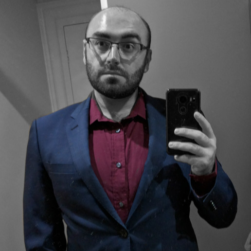 Jamie is a caucasian male in his mid 30s. The image is monochrome but has been edited to include the colour of the suit and shirt that he is wearing - navy blue and red, respectively. He has no hair on his head, but has a stubbly beard and square glasses.